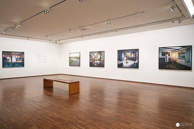 <p><a href="https://galleries.seirer-photography.com">seirer-photography</a><br />This installation view captures a gallery room at the Albertina's Gregory Crewdson retrospective, featuring five large-format chromogenic prints mounted against pristine white walls under precise track lighting. The photographs display Crewdson's characteristic suburban tableaux with their cinematic staging and artificially enhanced lighting, while a simple wooden bench positioned in the center of the polished hardwood floor provides contemplative viewing space for visitors to engage with the densely detailed, psychologically charged scenes. The neutral gallery environment allows the saturated colors and meticulous compositions of Crewdson's work to command full attention, demonstrating the museum's commitment to presenting these elaborately constructed photographic narratives as fine art objects. (Image 24 of 31)