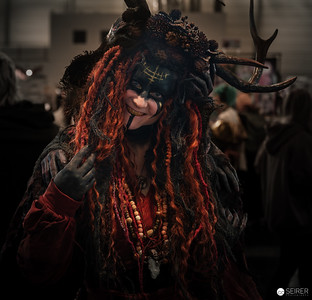 <p><a href="https://galleries.seirer-photography.com">seirer-photography</a><br />A cosplayer embodies a folkloric forest deity or shamanic figure, wearing an ornate headdress adorned with natural antlers, textured bark-like elements, and cascading auburn dreadlocks decorated with beads and talismans. The mask features dark, weathered surfaces with subtle embellishments, while dramatic side lighting emphasizes the sculptural quality of the costume against the softly blurred convention floor environment. Photographed at VIECC 2025 in Vienna, the composition centers the performer's theatrical gesture—hand raised to the masked face—creating an enigmatic portrait that blurs the boundary between cosplay craftsmanship and performance art, exemplifying the elaborate character interpretations prevalent at contemporary fan conventions. (Image 32 of 162)