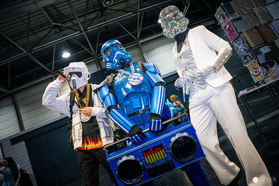 <p><a href="https://galleries.seirer-photography.com">seirer-photography</a><br />Three costumed figures occupy the central exhibition space beneath industrial ceiling infrastructure at VIECC 2025, Vienna's Comic Con event. The composition features a Stormtrooper costume (left), a chrome blue armored figure holding a decorated boom box (center), and a figure in white suit with crystalline mask (right), arranged in a triangular formation that creates visual depth through their varied heights and metallic surface reflections. The lighting emphasizes the contrast between the matte white armor and highly reflective blue chrome finish, while the boom box's decorative elements add a hip-hop cultural reference to this Star Wars-inspired ensemble, exemplifying the creative fusion of pop culture fandoms characteristic of contemporary cosplay communities. (Image 72 of 162)